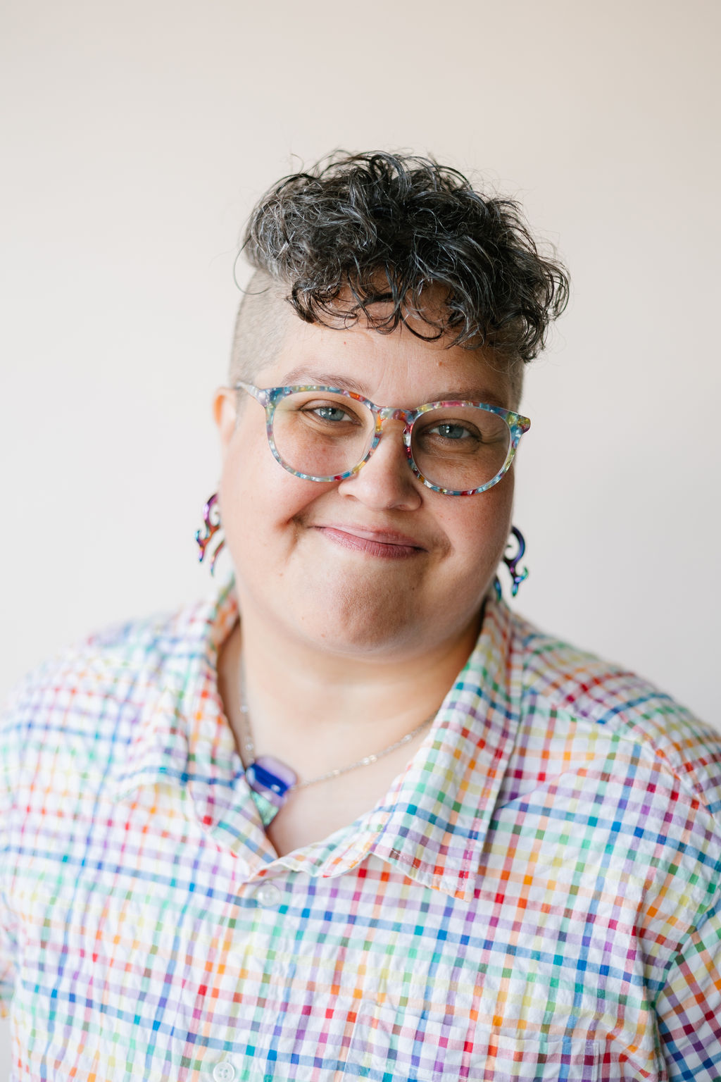Portrait of Lior Effinger-Weintraub, a light-brown skinned human with grey eyes, a shaved undercut and curly greying brown hair. Lior is wearing colorful glasses, earrings, and a buttonup shirt with rainbow colors.